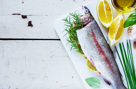 Top view of old wooden table with fresh raw rainbow trout on white marble board with lemon, herbs and spices, copy space. Seafood cooking background, preparation.の写真素材
