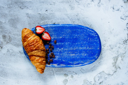 Blue chopping board with freshly baked croissant, strawberries and coffee beans on concrete textured backdrop, top view, space for text. Breakfast or dessert setの写真素材