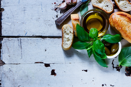 Fresh baguette cut in slices with basil, garlic, spices and green olives over old white wooden backdrop, top view, copy space.の写真素材