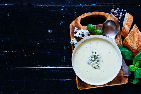Healthy home cooked cream soup of yellow lentils, served with blue cheese, fresh basil and bread slices on old chopping board over grunge wooden background. Dark rustic style. Top view, copy space.の写真素材
