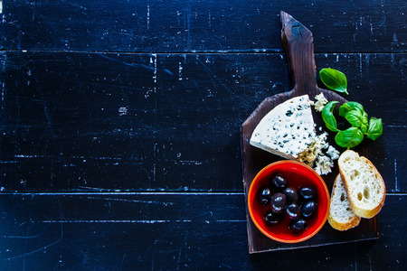Fresh blue cheese with olives, basil and bread slices on dark wooden chopping board over old grunge background, top view, copy space.の写真素材