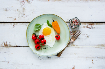 Top view of breakfast set. Fried egg, roasted cherry tomatoes, fresh basil and spices on ceramic plate over white wooden backdrop, horizontal composition. Healthy, clean eating, dieting food conceptの写真素材