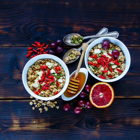 Delicious oatmeal for breakfast, granola in bowls, dried berries, seeds, honey, bloody orange, grapes over dark wooden background, square crop. Clean eating, vegan, vegetarian, detox and dieting conceptの写真素材