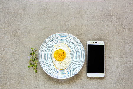 Breakfast fried egg in plate and smartphone flat-lay. Home rest break still life on light stone background, top view.の写真素材