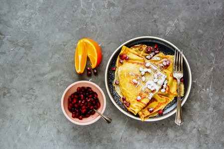 Sweet crepes topped with pomegranate and sugar on plate flat lay. Homemade thin pancakes and fruits