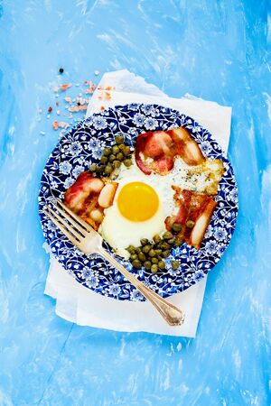English breakfast in plate with fried egg and bacon on blue background flat layの写真素材