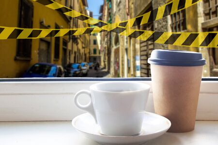 Coffee cup and take away coffee on the windowsill with an Italian landscape outside the window. Barrier tape, quarantine, isolation, coronavirus conceptの写真素材