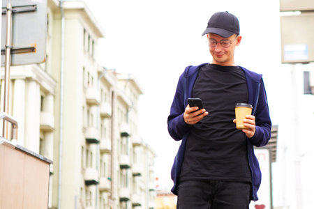 Young man walking through city street and holding takeaway coffee and phone
