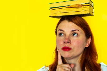 Redhead girl with books on yellow background. Teacher is holding books on her head. Back to school conceptの写真素材