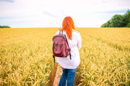 Ginger girl with backpack on the road in a wheat field. Summer time conceptの写真素材
