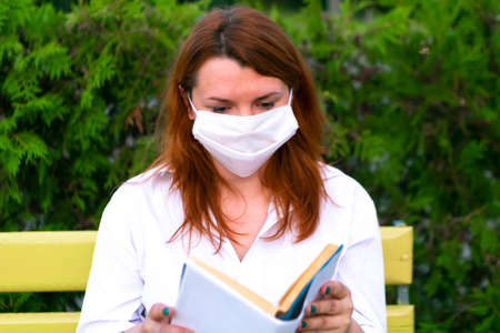 Girl in face mask sits on the bench and read book in the park, coronavirus conceptの写真素材