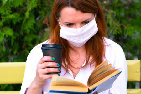 Girl in face mask with coffee cup sits on the bench and read book in the park, coronavirus conceptの写真素材