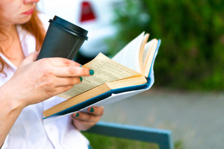 Woman with coffee cup is reading a book in the park, closeup