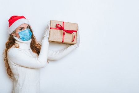 Woman in face mask and Santa hat holding gift box with red ribbon. Coronavirus Christmas concept. Copy space for the textの写真素材