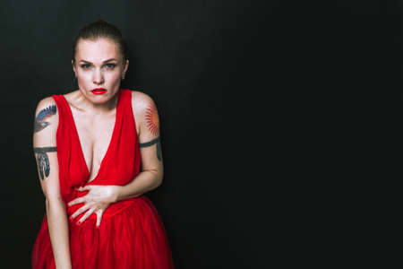 Fashion model in red beauty dress. Sexy woman with tattoos is posing in evening dress. Studio shot with black isolated backgroundの写真素材