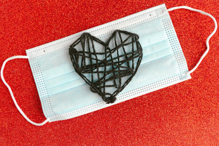 Black knitted heart and medical mask on red glitter background. Valentine's Day during the coronavirus pandemic. Happy Valentine's Day 2021.の写真素材