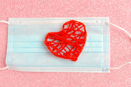 Red knitted heart and medical mask on pink glitter background. Valentine's Day during the...