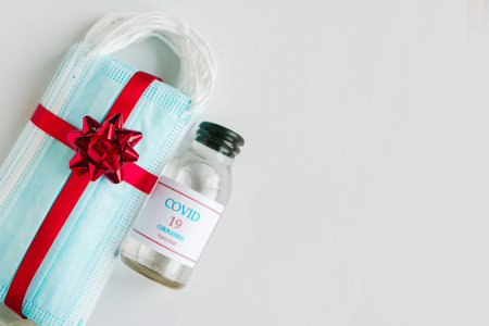 Stack of medical masks with gift bow and covid-19 vaccine as a present on white background....