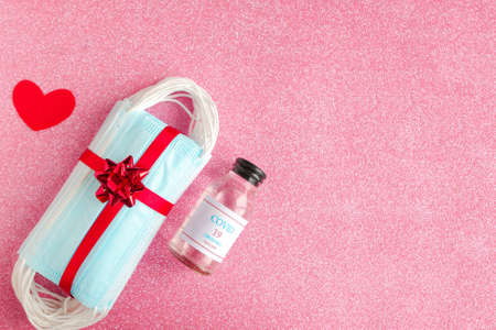 Stack of medical masks with gift bow and covid-19 vaccine as a present on pink glitter background. Valentine's Day during the coronavirus pandemic. Copy space for the textの写真素材