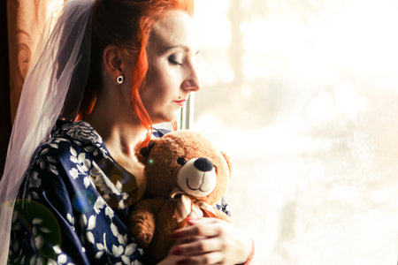 Portrait of young bride in wedding dress and veil. Upset girl is standing near the window and holding a teddy bearの写真素材