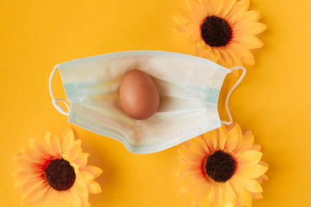 Chicken egg in face medical mask and yellow flowers. Easter holiday during the coronavirus pandemic