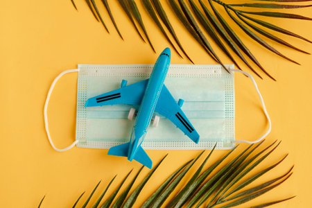 Toy airplane, medical mask and palm tree branches on yellow background. Coronavirus travel. Summer...
