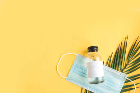 Face medical mask, hand sanitizer and tropical green leaves on bright yellow background minimal. Coronavirus travel. Summer holidays during the pandemic concept. copy spaceの写真素材