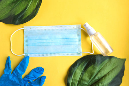 Face medical mask, hand sanitizer and palm three leaves on bright yellow background. Coronavirus...