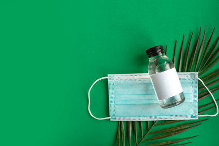 Face medical mask, sanitizer bottle and tropical leafs on green background minimal. travel. Summer...