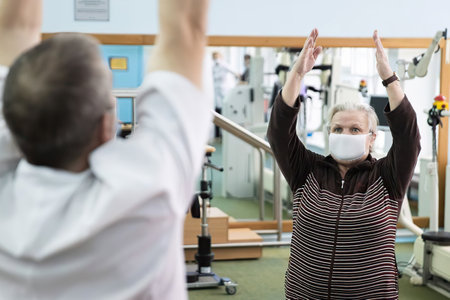 Minsk Belarus â February 10 2021: Rehabilitation after coronavirus infection. Elderly woman in face medical mask do restorative exercises with a doctor in a gym during covid-19 pandemicのeditorial素材