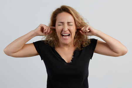 Emotional beautiful young adult woman portrait. Studio shoot. Isolated on White Background. The woman screams, showing perfect teeth, screwing up her eyes, shutting her ears with her fingers.の写真素材