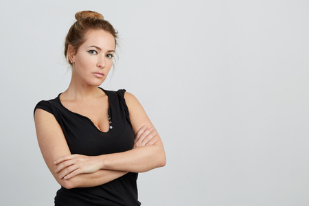 Emotional beautiful young adult woman portrait. Looking at Camera. Studio shoot. Isolated on White Background. A woman in a black T-shirt with a deep neckline crossed her arms over her chest.の写真素材