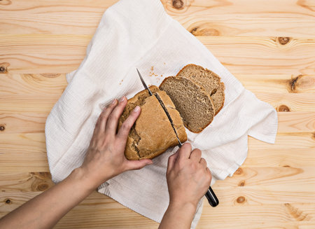 Female hands cut freshly baked homemade rye bread with a knife into slices on a white towel,...
