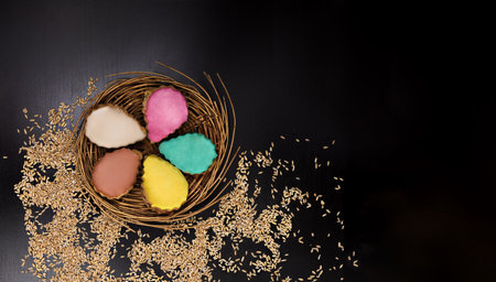 Multi-colored cakes (turquoise, beige, pink, yellow and caramel) served on a wicker dish around scattered grains on a black background, horizontal, copyspace on the right, top view, flat layの写真素材