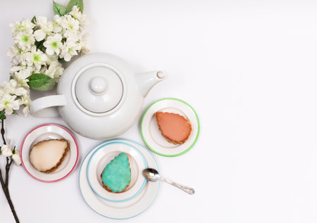 Multi-colored cake served on saucers around a white teapot on a white background with flowering branches, top view, flat lay, isolated on white, copyspase on the right, horizontalの写真素材