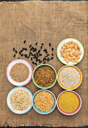 Portions of various cereals - wheat, peas, buckwheat, millet, oatmeal, barley in a colored dish against a background of burlap and scattered sunflower seeds and flakes, top view, flat lay, close up, copy space, verticalの写真素材