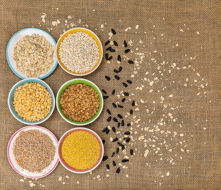 Portions of various cereals - wheat, peas, buckwheat, millet, oatmeal, barley in a colored dish against a background of burlap and scattered sunflower seeds and flakes, top view, flat lay, close up, copy spaceの写真素材