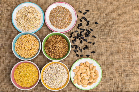 Portions of various cereals - wheat, peas, buckwheat, millet, oatmeal, barley in a colored dish against a background of burlap and scattered sunflower seeds and flakes, top view, flat lay, close up, copy spaceの写真素材
