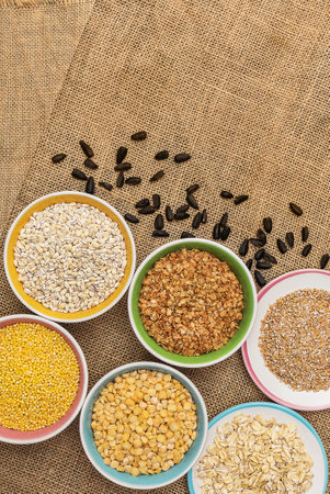 Portions of various cereals - wheat, peas, buckwheat, millet, oatmeal, barley in a colored dish against a background of burlap and scattered sunflower seeds, top view, flat lay, close up, copy space, verticalの写真素材