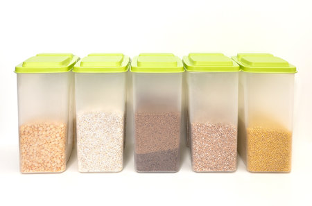 Various cereals - wheat, buckwheat, millet, barley in closed plastic transparent containers for bulk products, standing in a row, on a white background, copy space, vertical, front view, shallow depth of fieldの写真素材