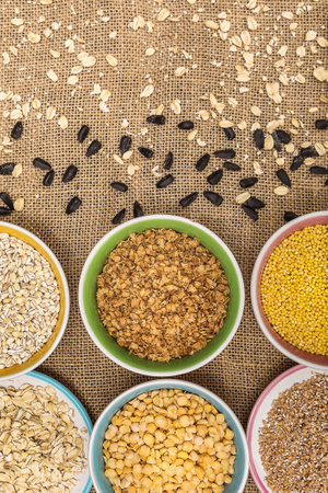 Portions of various cereals - wheat, peas, buckwheat, millet, oatmeal, barley in a colored dish against a background of burlap and scattered sunflower seeds and flakes, vertical, top view, close upの写真素材