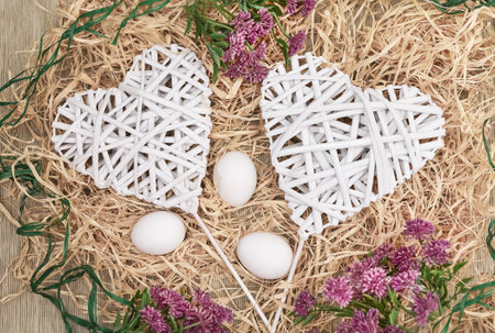 Decorative braided hearts on a natural raffia background, surrounded by clover flowers and eggs, close-up, top view; white, purple and green on straw yellowの写真素材