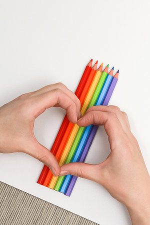 Female hands show the shape of a heart on a background of a rainbow made of colored pencils and a white sheet of paper diagonally, close-up, copy spaceの写真素材