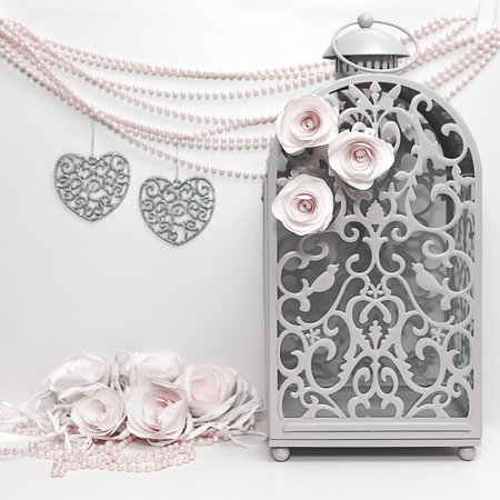 Refined ornamented lantern, decorated with white roses, against a background of decor with two openwork figures of hearts and pearl beads in pale pink tonesの写真素材