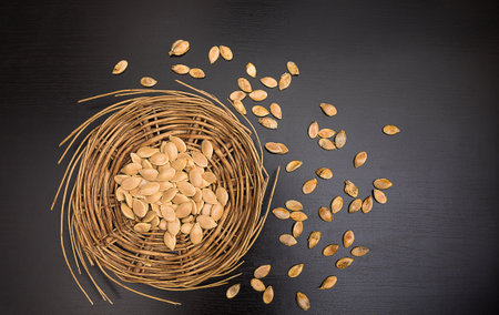 A handful of healthy pumpkin seeds peeled on a round wicker tray and scattered on a black table, horizontal, top view, close upの写真素材