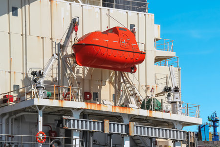 A red lifeboat, securely attached to a large ship by a robust hook, contrasts with the white ship and clear blue sky at a bustling port, promising a safe and adventurous journey.の写真素材