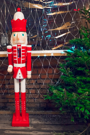 A Nutcracker in a red coat and hat next to a Christmas tree with red and white ornaments, topped with a star. The tree has twinkling white lights against a white background with a brick wall texture.の写真素材