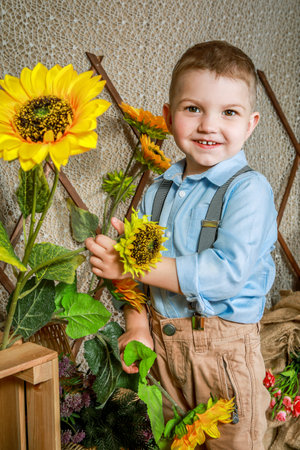 A cheerful young boy stands next to a crate filled with sunflowers. He is wearing a blue button-up shirt, brown pants, and suspenders. The background is a rustic wooden fence.の写真素材