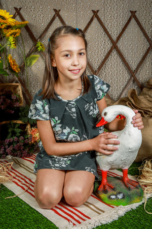 A smiling girl in a floral dress with a ponytail, sitting with a goose on a blanket. Rustic background with hay and wooden fence.の写真素材