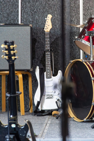 A white electric guitar leans against an amplifier next to a drum kit set up on stage. The space is...